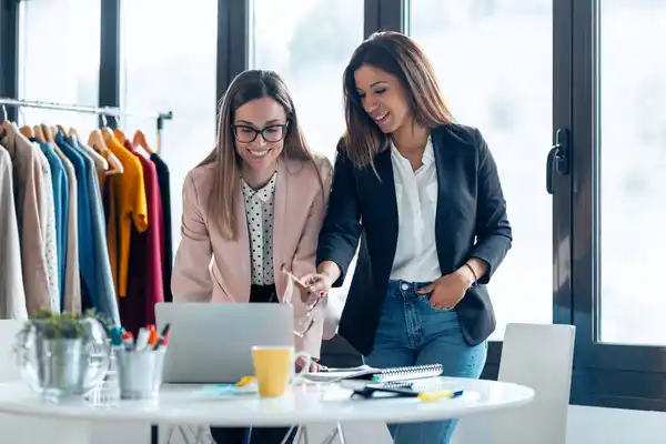 Dos mujeres emprendedoras analizan plan de dirección y gestión empresarial en laptop en una oficina con ropa en un estante al fondo.
